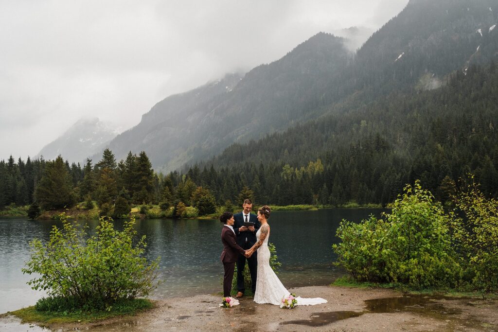 Brides hold hands during their LGBTQ elopement ceremony by an alpine lake