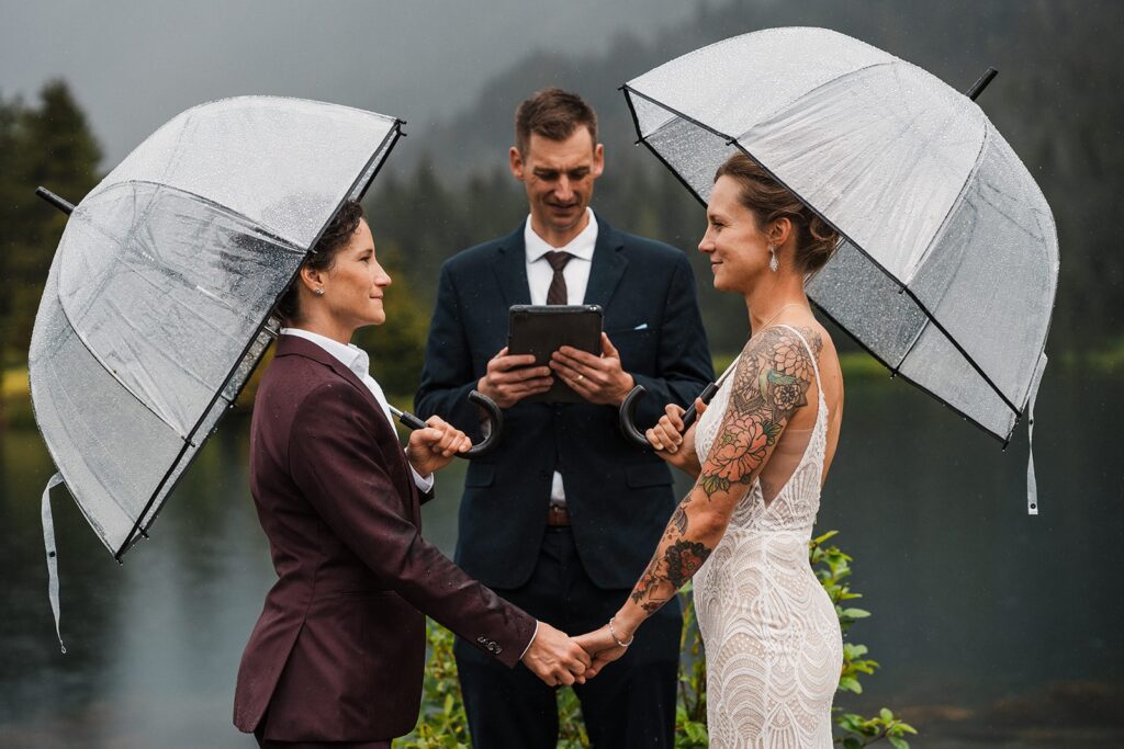 Brides hold hands during their rainy wedding ceremony by an alpine lake in Snoqualmie, WA