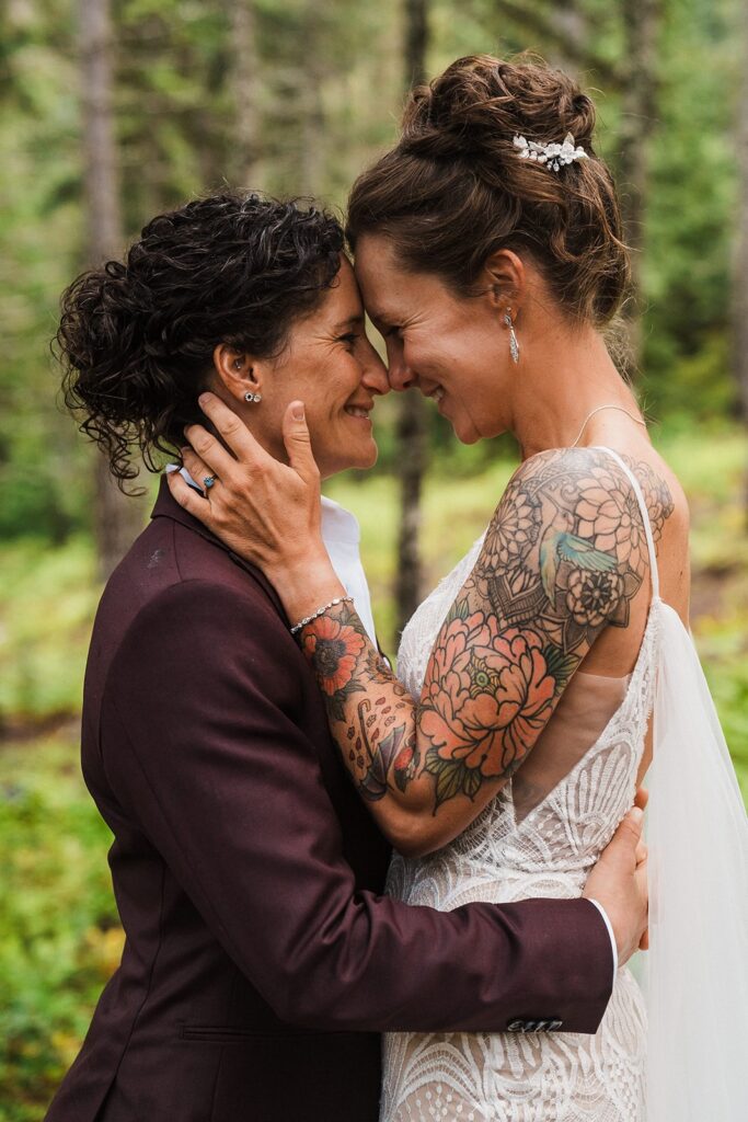 Two brides stand forehead to forehead in the forest during their LGBTQ wedding 