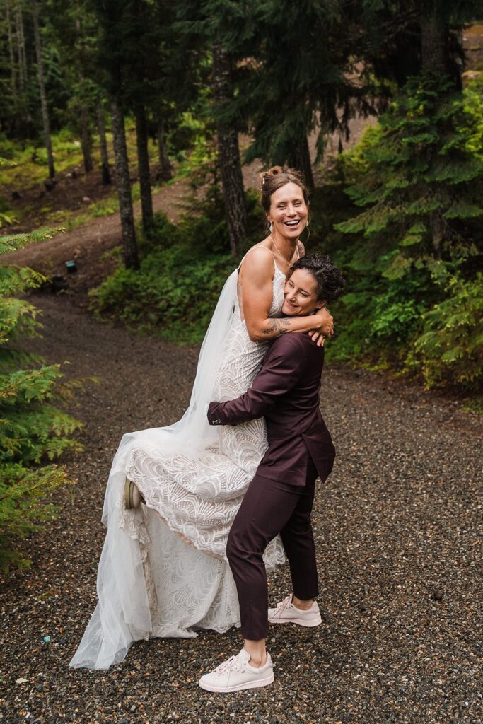 Bride picks up the other bride on a forest trail during their Washington wedding 