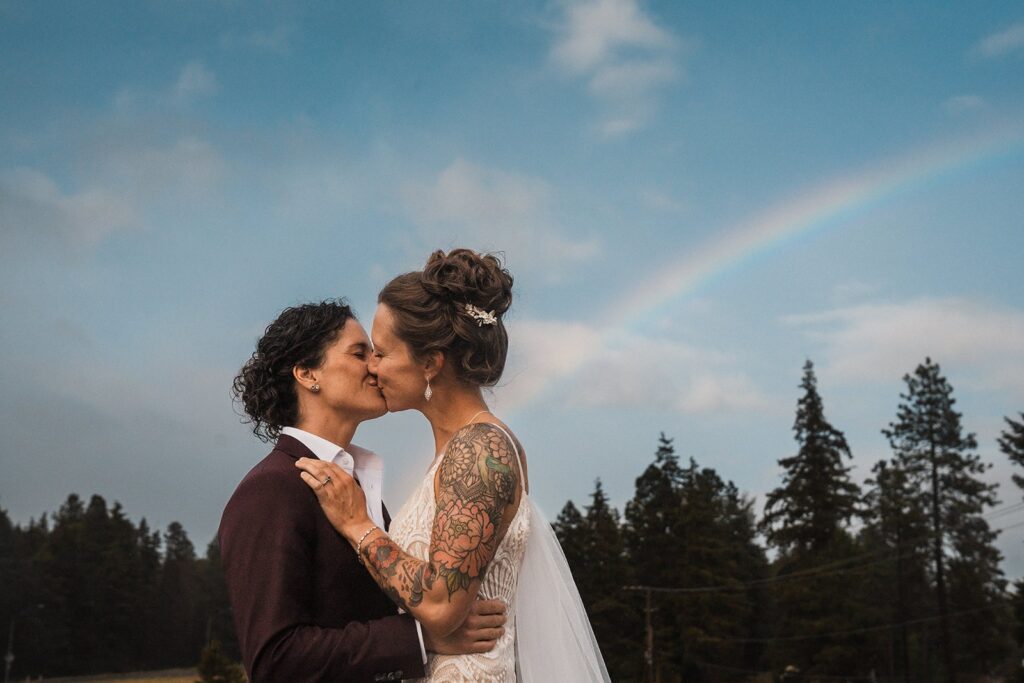 Brides kiss in front of a rainbow in the sky during their LGBTQ elopement 