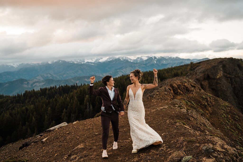 Brides cheer while walking along a mountain trail during their Washington wedding during pride month