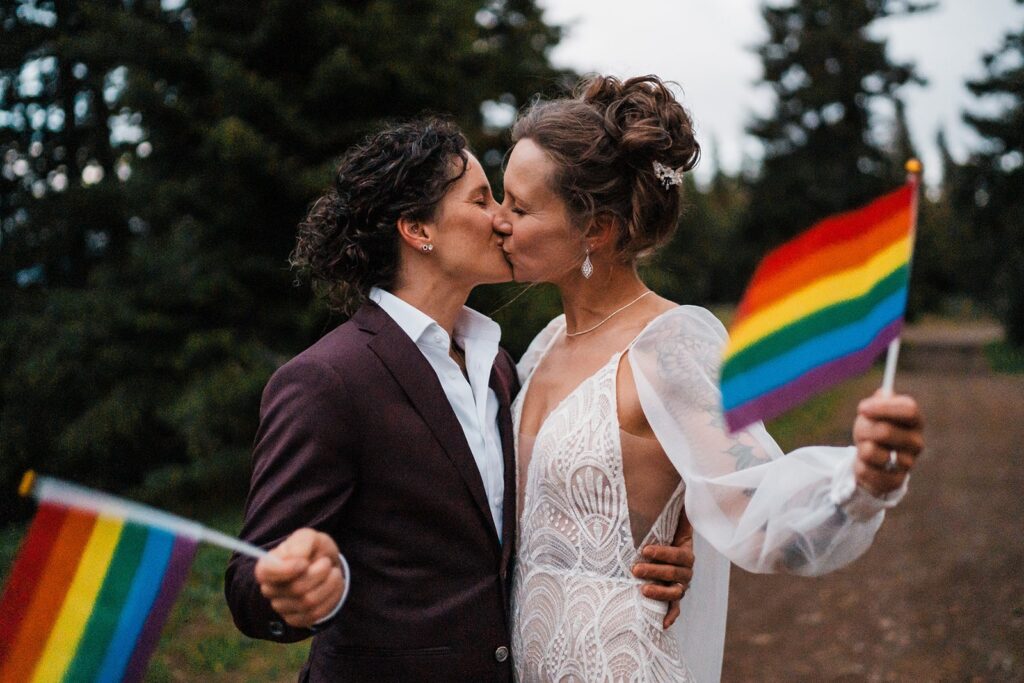 Brides wave pride flags while they kiss at their LGBTQ elopement