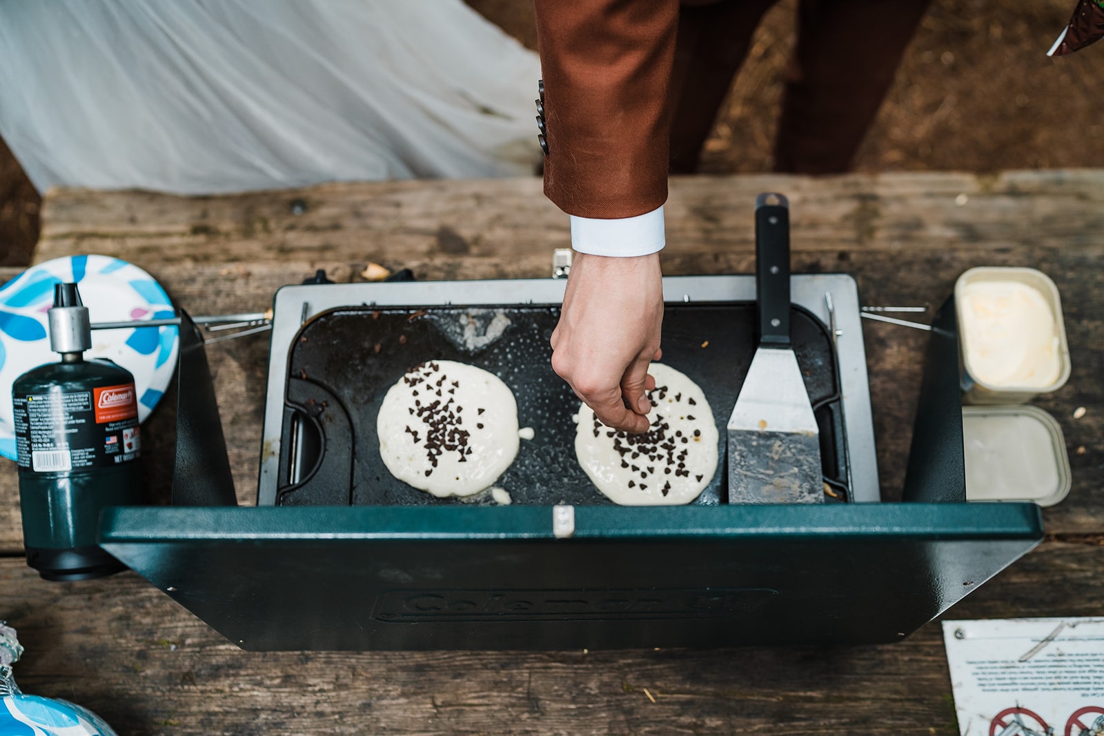 Groom sprinkles chocolate chips on cookstove pancakes at a day use area