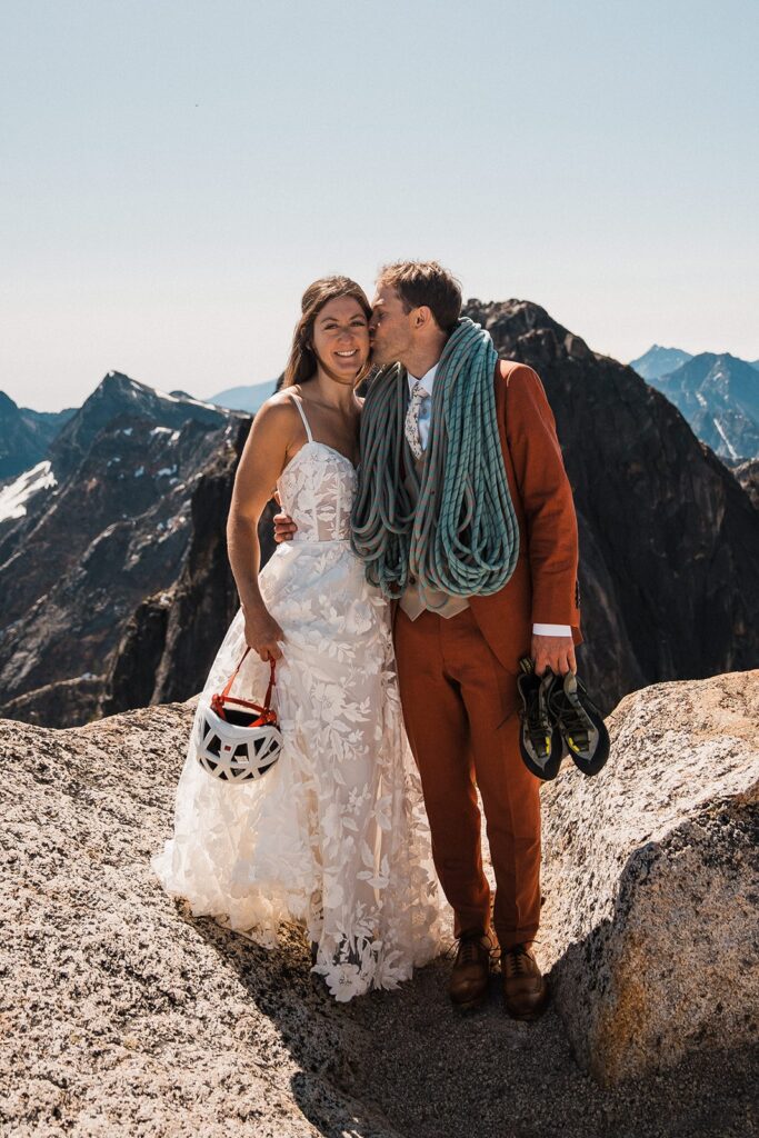 Bride and groom hold climbing gear in their wedding attire on top of a mountain peak in the North Cascades