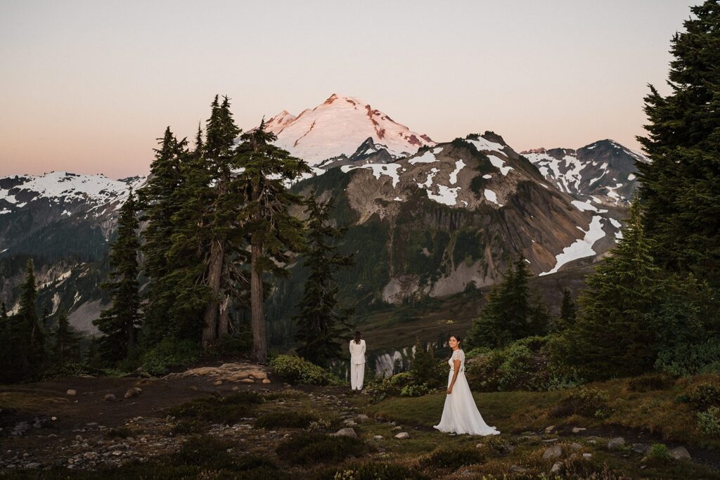 Bride walks up behind another bride for their first look on a mountain trail in the North Cascades 