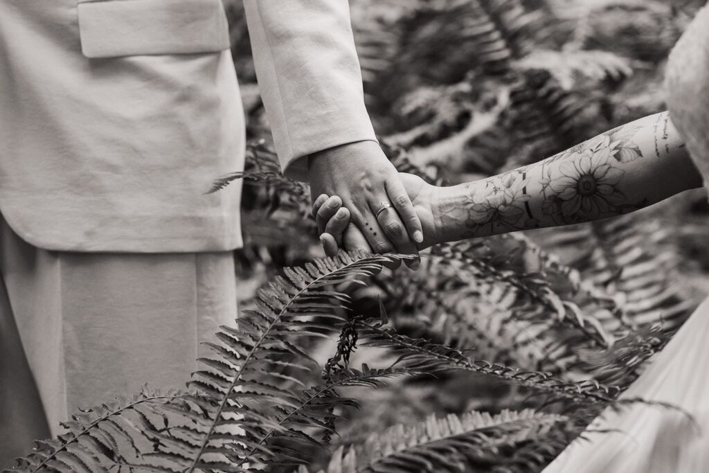 Brides hold hands as they walk through the fern-filled forest during their summer elopement 