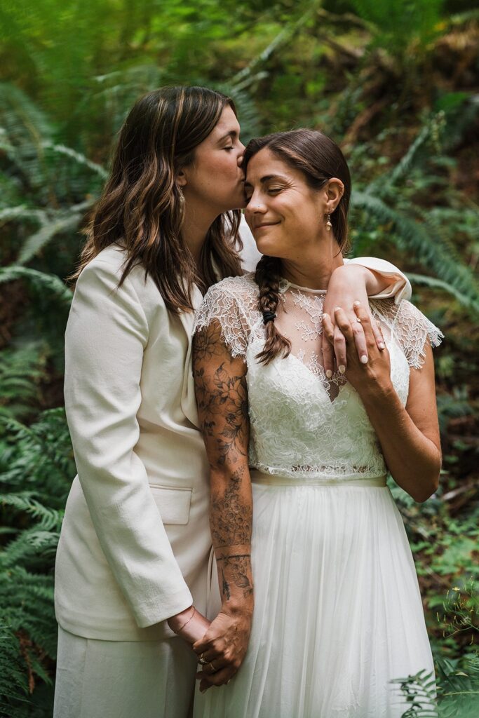 Bride kisses her bride on the forehead during their forest summer elopement in the North Cascades