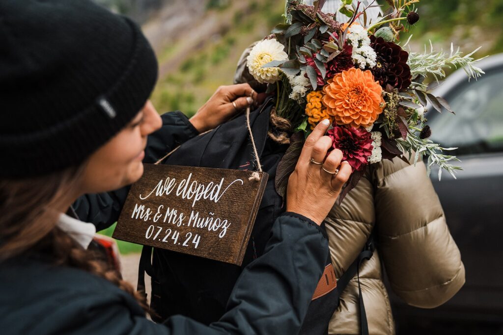 Brides adjust their hiking backpack and flowers before hiking a trail in the North Cascades during their summer elopement 