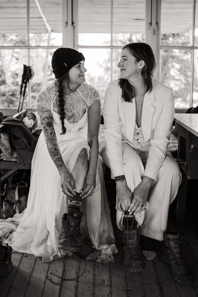 Brides smile at each other while lacing up their hiking boots in a mountain hut in the North Cascades