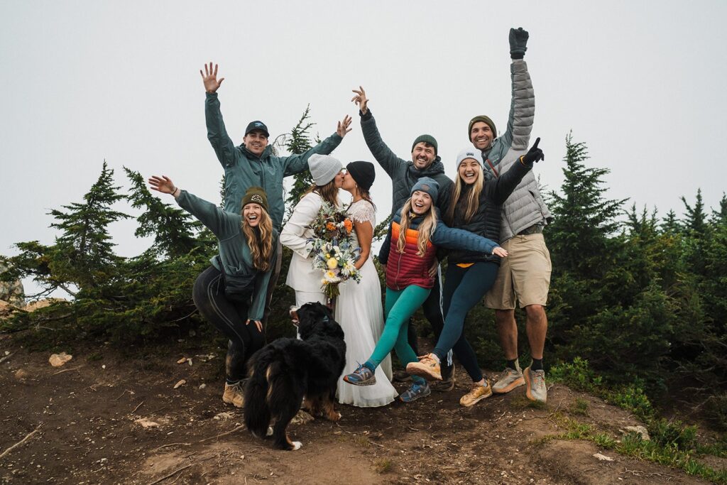 Friends cheer while brides kiss on a mountain trail during their summer elopement 