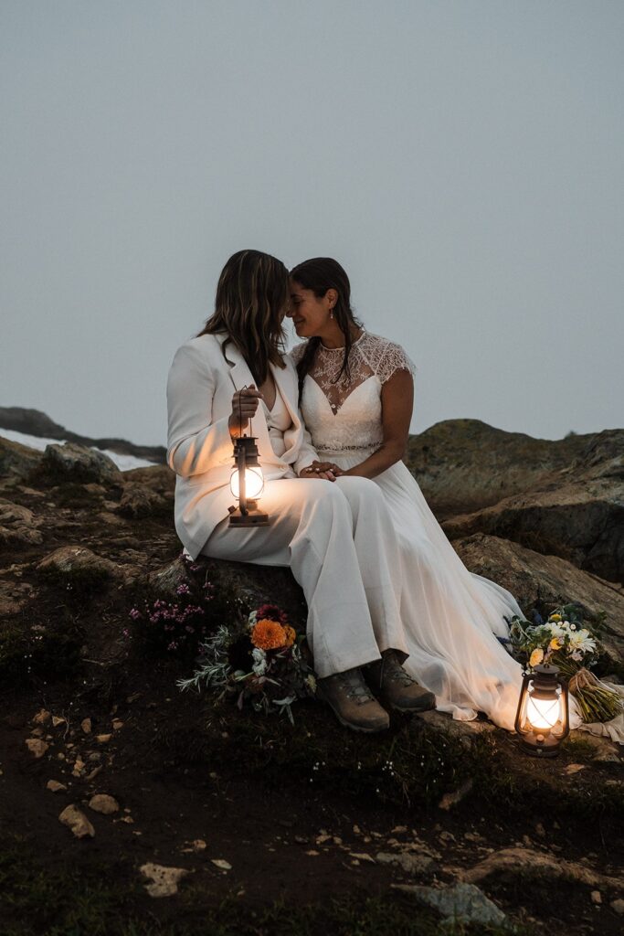 Brides sit on a mountain trail while holding lanterns during blue hour