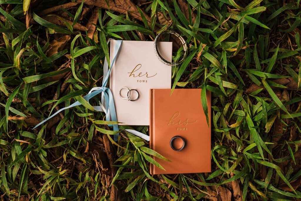 Pink and orange vow books lay in the grass with wedding bands laying on top 