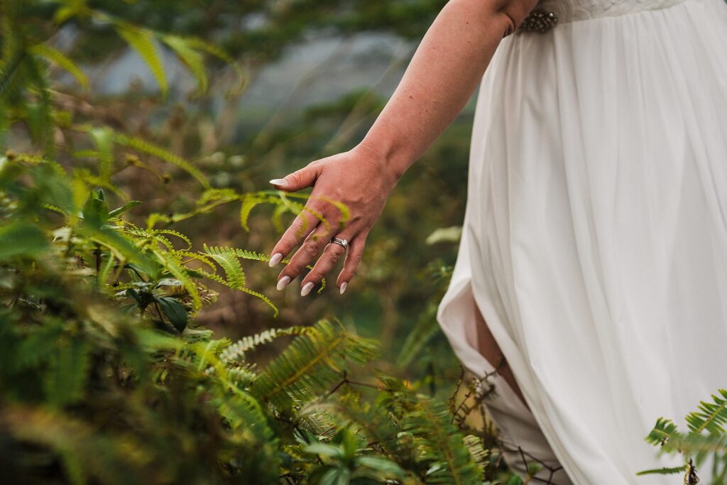 Bride runs her hands along the ferns while walking a forest trail at her elopement in Kauai 