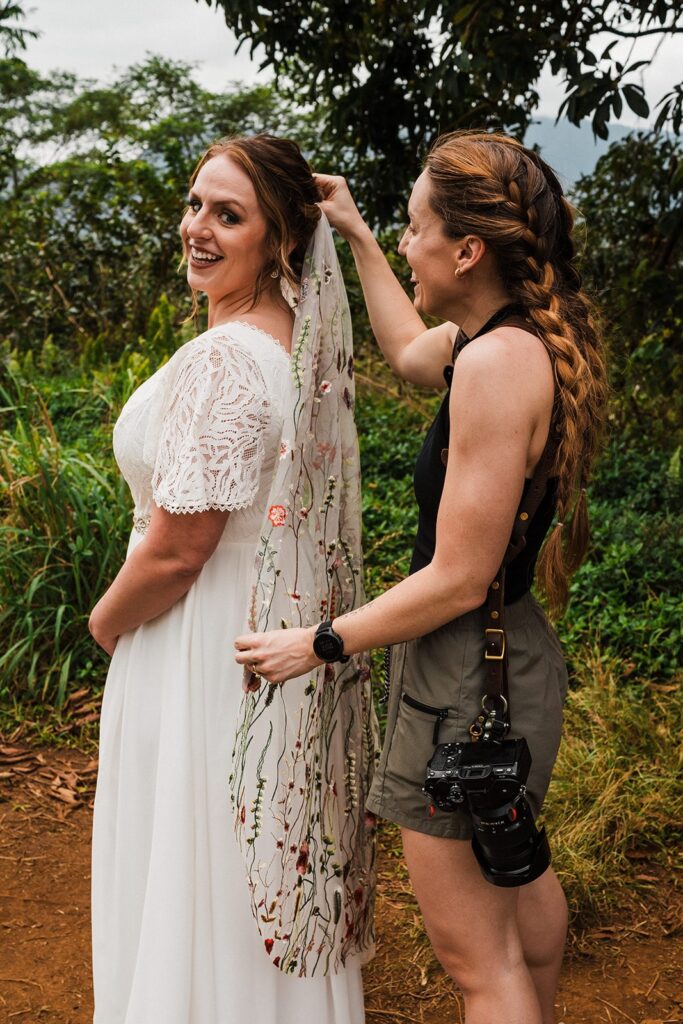 Kauai elopement photographer helps bride put on her embroidered floral veil at her outdoor elopement in Kauai 