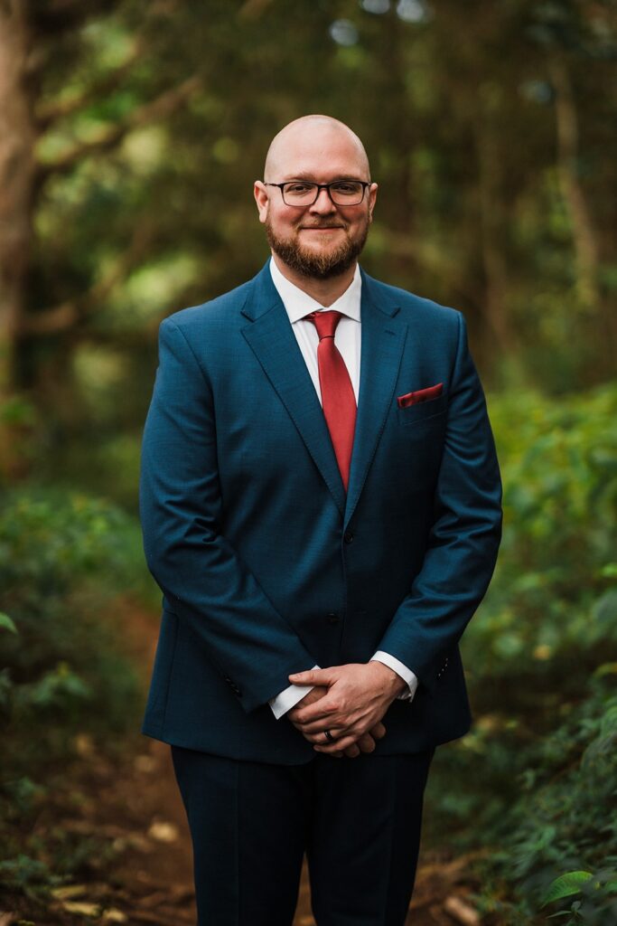 Groom wears a blue suit and red tie in the forest at his Kauai elopement 