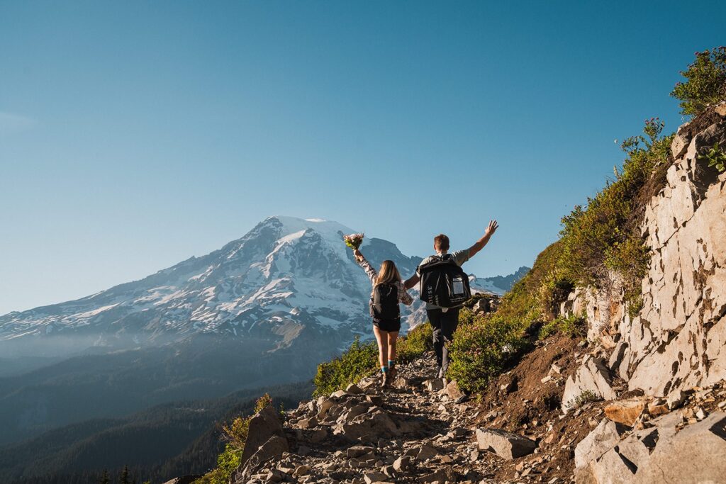 Bride and groom raise their hands in the air while hiking a mountain trail toward Mount Rainier 