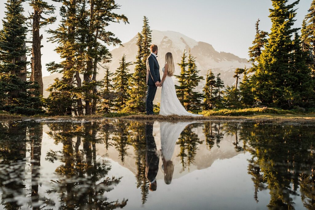 Bride and groom hold hands by a tarn while looking out at Mt Rainier in the distance 