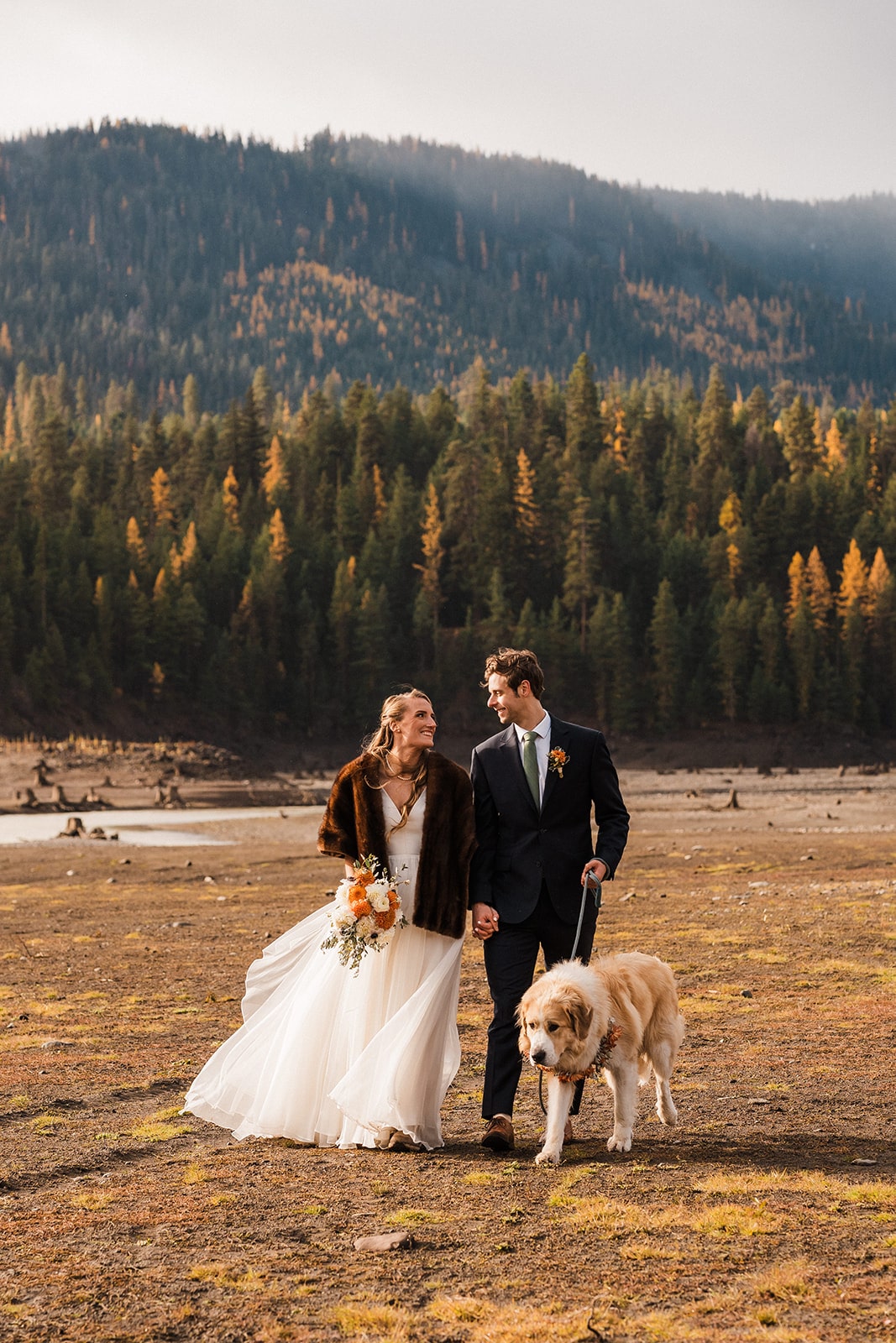 Bride and groom hold hands while walking through a field with larch trees dotting the mountains behind them