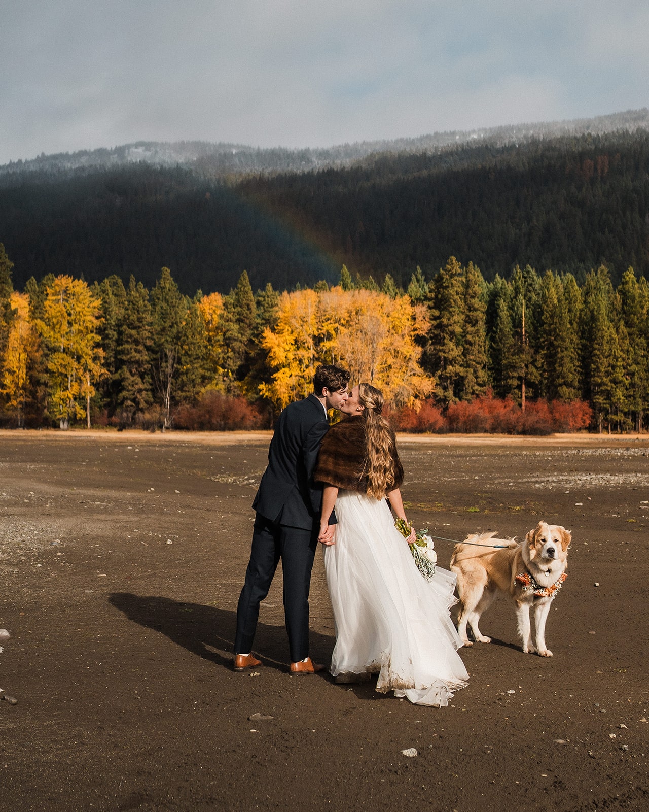 Bride and groom kiss and hold hands while walking through a field with larch trees dotting the mountains behind them