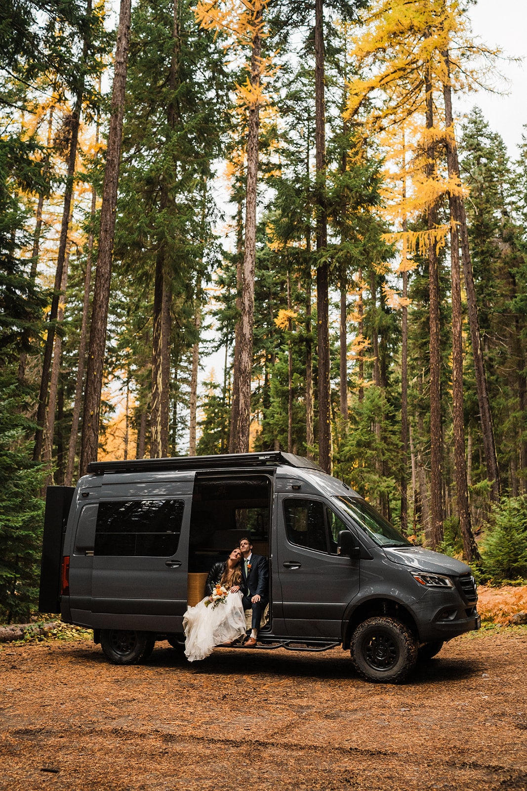 Bride and groom sit in their gray camper van in the forest before heading out on their fall wedding adventure hike