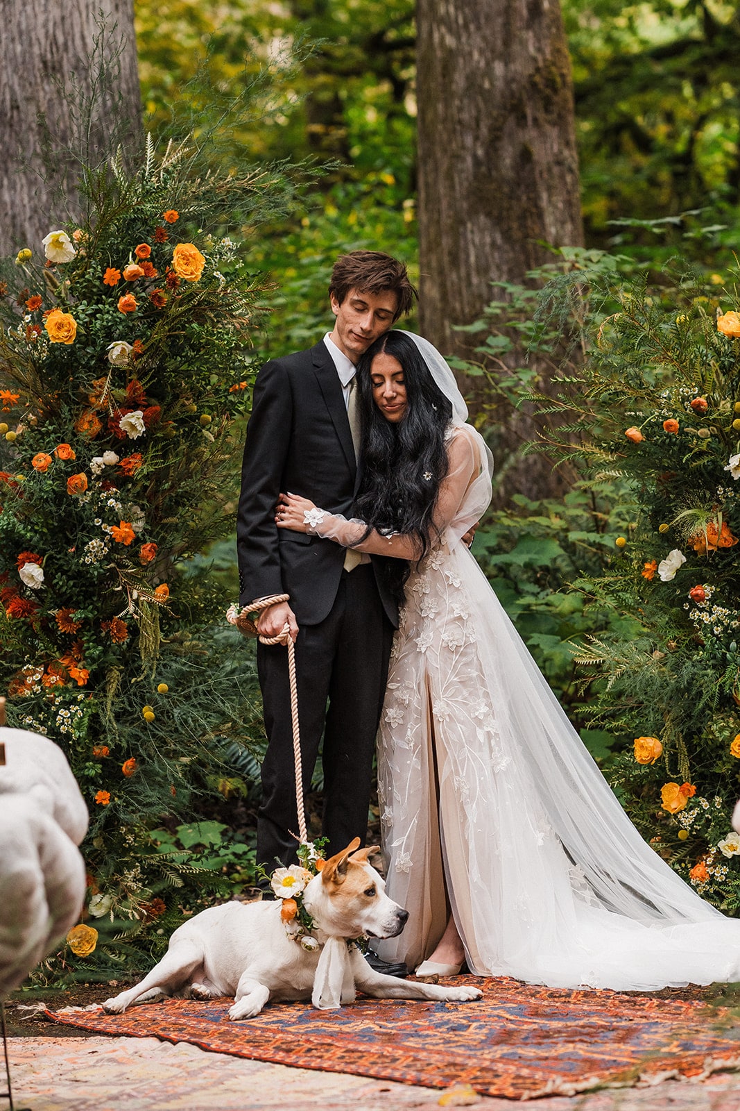 Bride and groom hug each other at their forest wedding while their dog lays on a rug at their feet