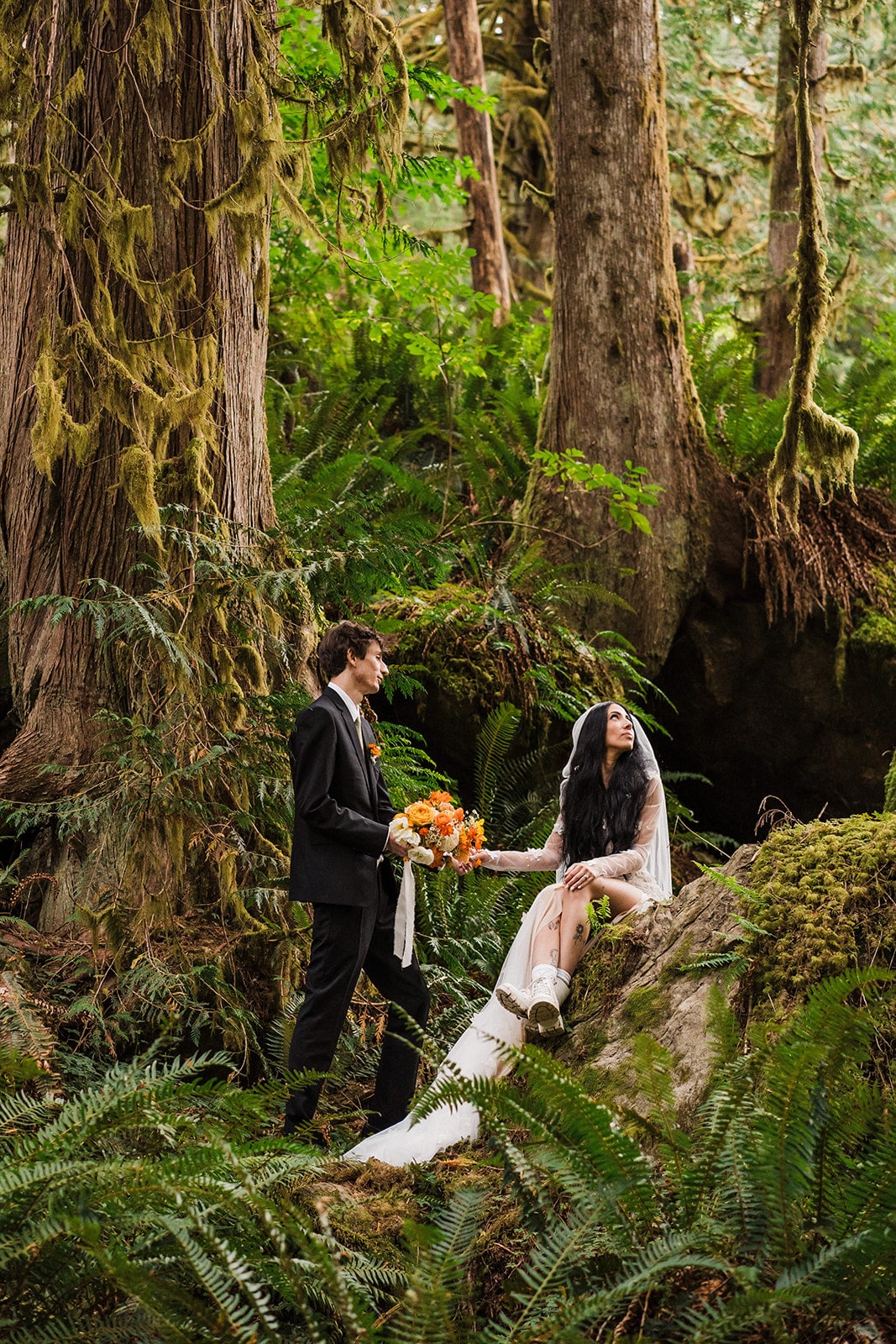 Bride sits on a mossy rock while holding groom's hand during their Mt Baker elopement in the forest 