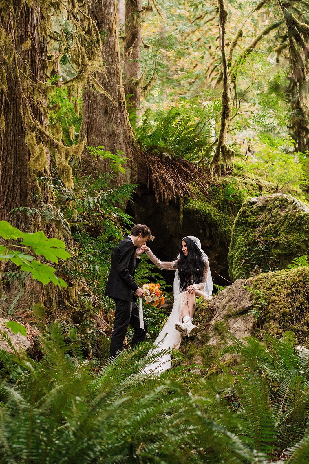 Groom kisses bride's hand while she sits on a mossy rock in the forest during their Mt Baker elopement photos 