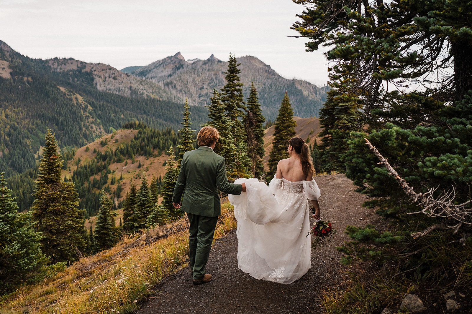 Groom holds bride's wedding dress train as they walk along a mountain trail during their national park elopement