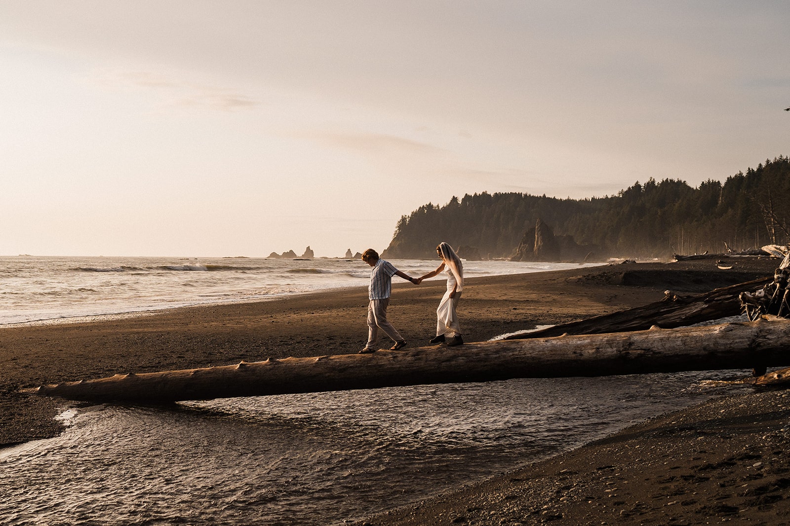 Bride and groom hold hands while walking across a fallen tree over a stream at Rialto Beach