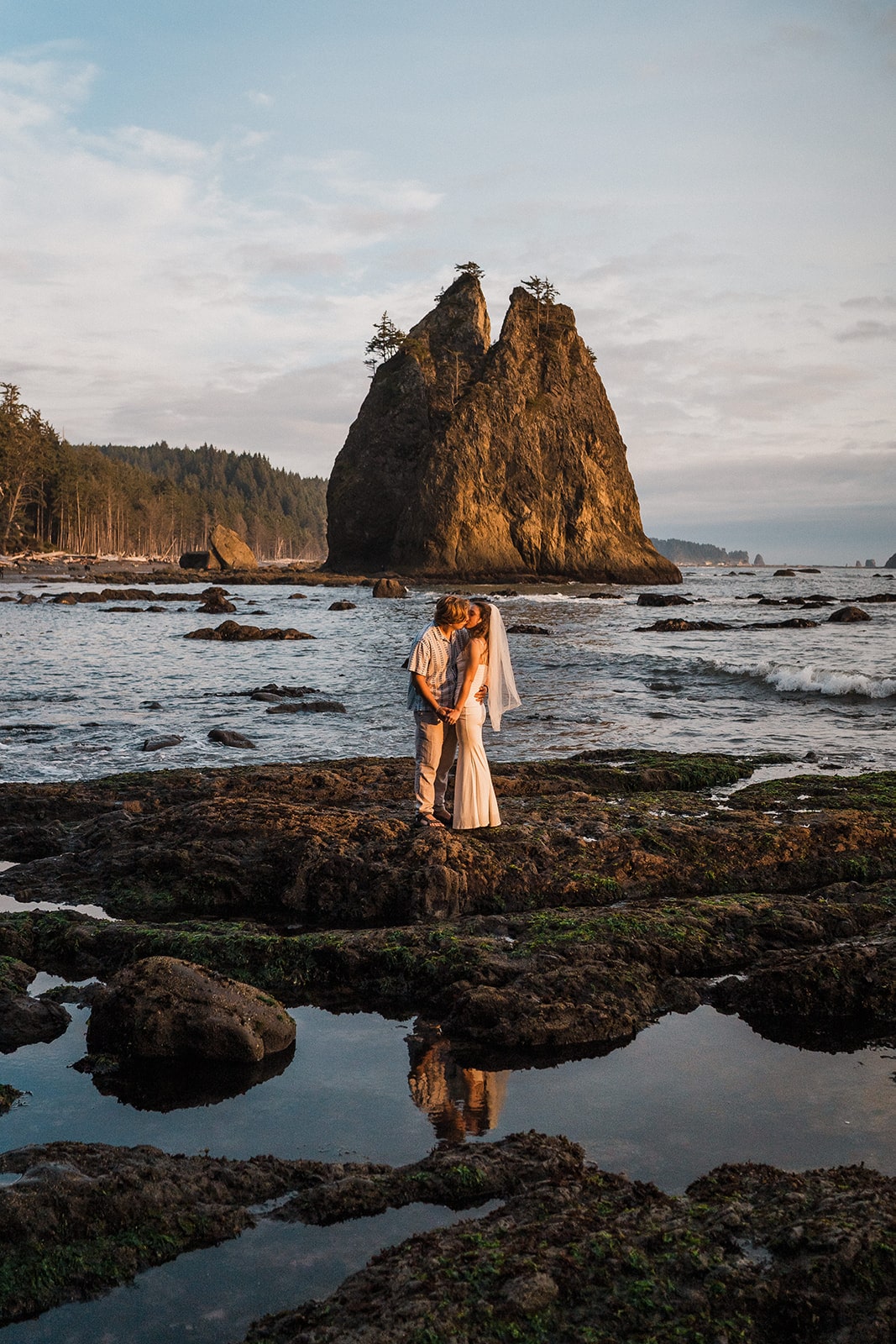 Bride and groom kiss while standing on moss covered rocks at Rialto Beach