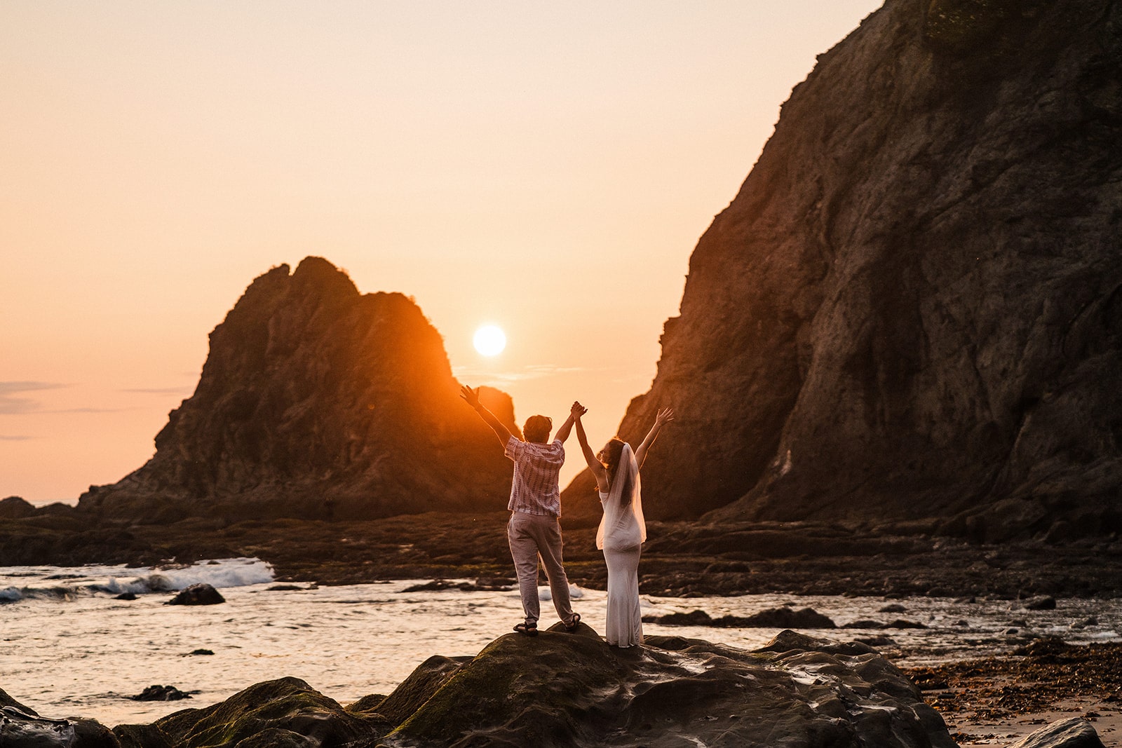 Bride and groom stand on a rock and cheer during their sunset adventure photos at Rialto Beach