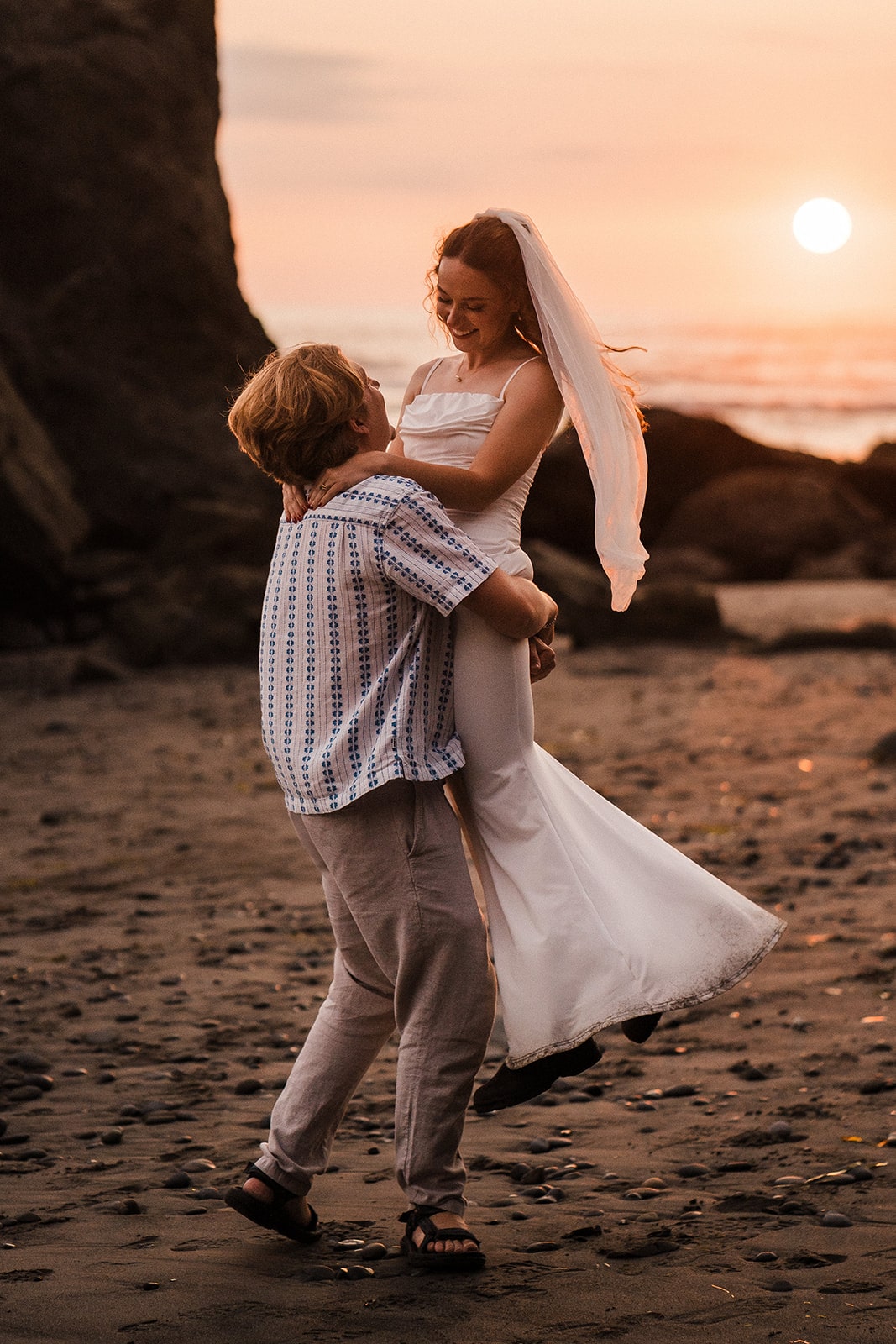 Groom lifts bride up and twirls her around during their sunset beach photos at Rialto Beach