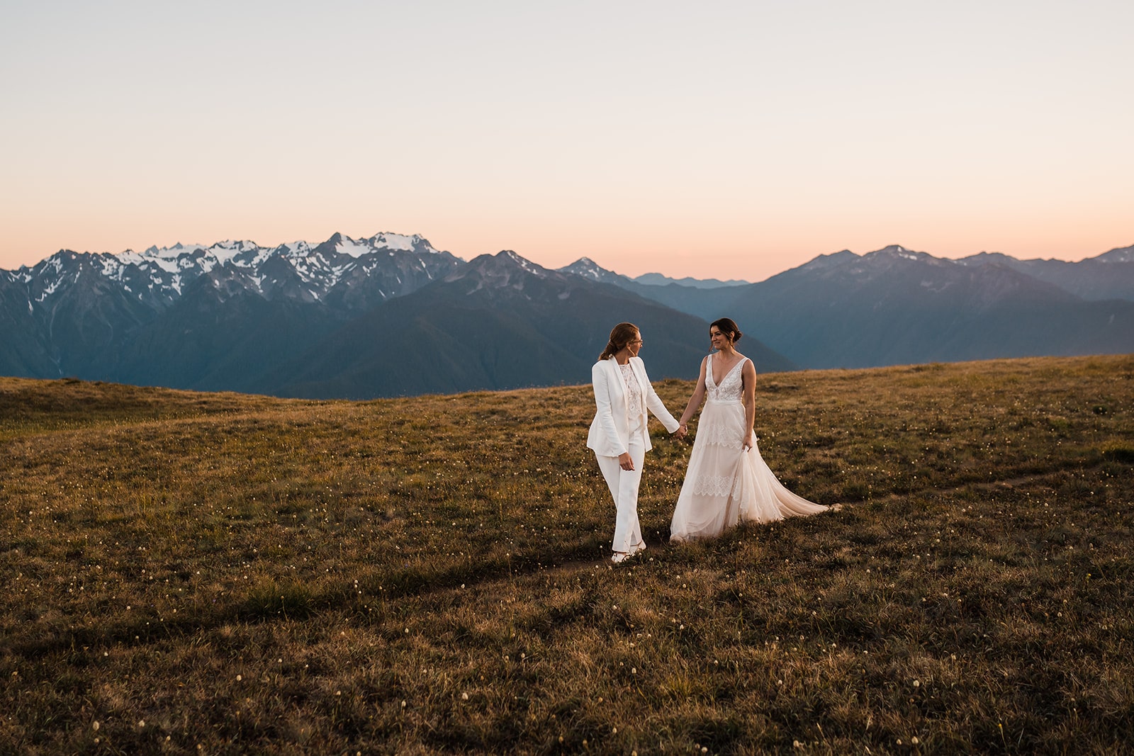 Two brides in elopement attire hold hands while walking across a mountain field during their adventure elopement 