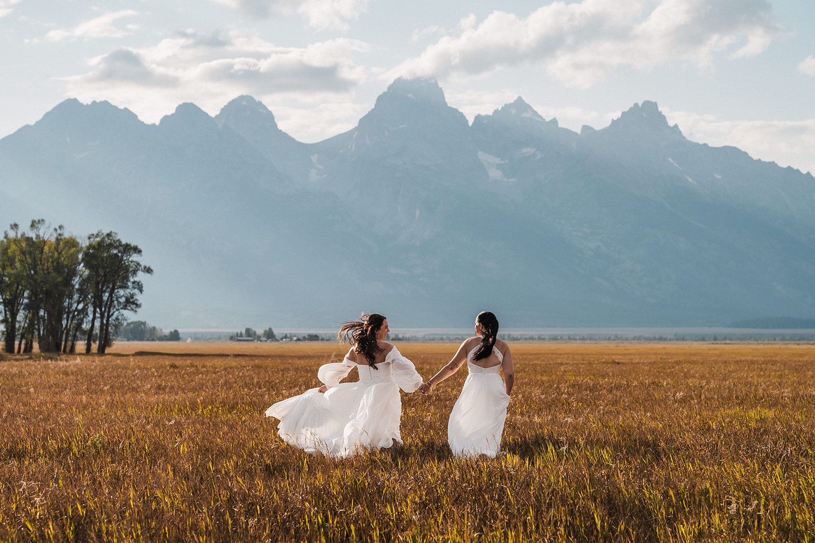 Two brides in white wedding dresses hold hands while running through a field during their adventure elopement