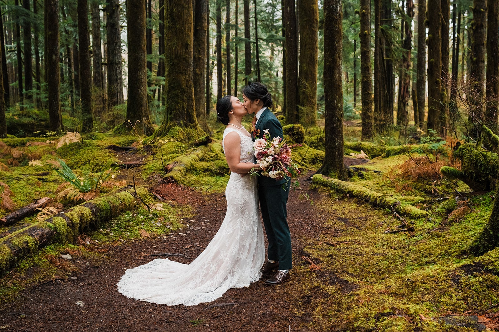 Two brides kiss in the forest while wearing a white elopement dress and green elopement suit 