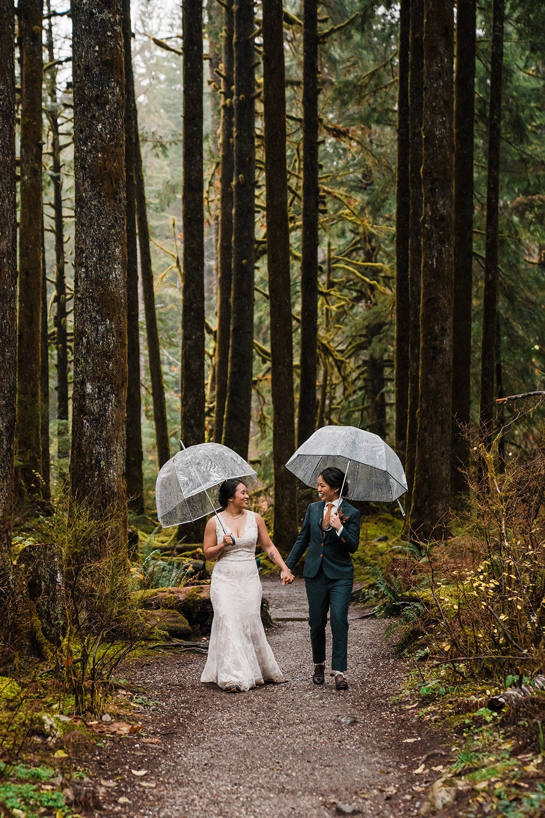 Brides hold hands while walking through the forest during their rainy elopement in Washington