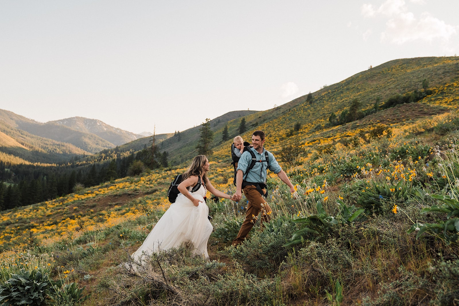 Bride and groom hold hands while hiking up a mountain trail with their son strapped to his dad's back in a baby carrier.