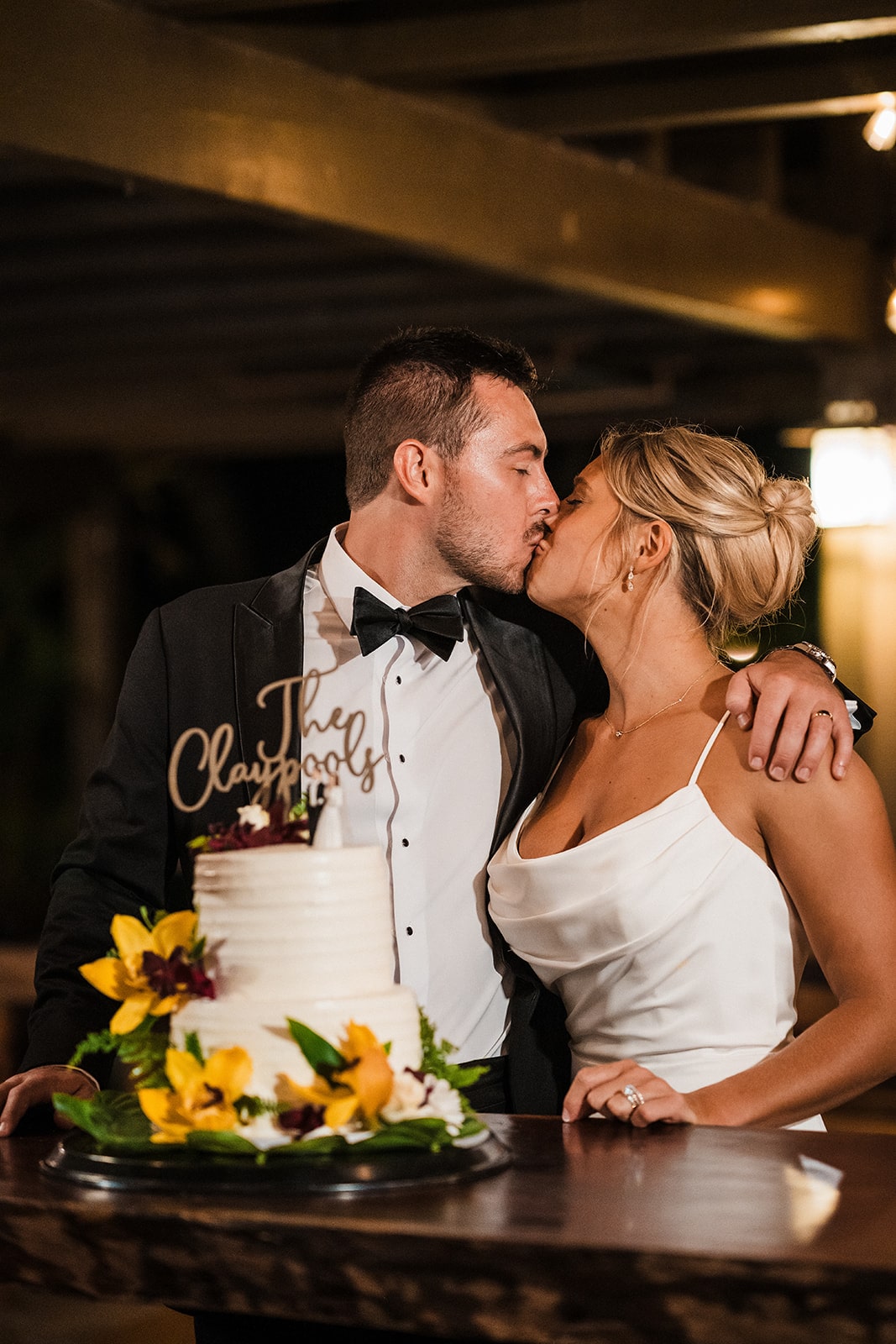 Bride and groom kiss in front of their two tier white wedding cake with yellow flowers and a custom topper.