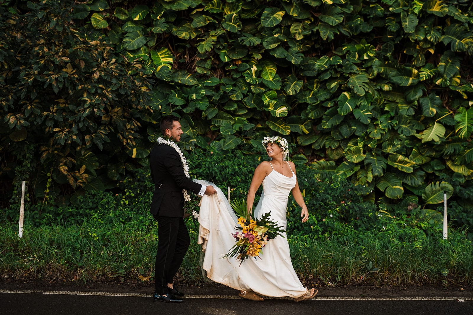 Groom carries bride's dress train while they walk along a road during their Kauai wedding.