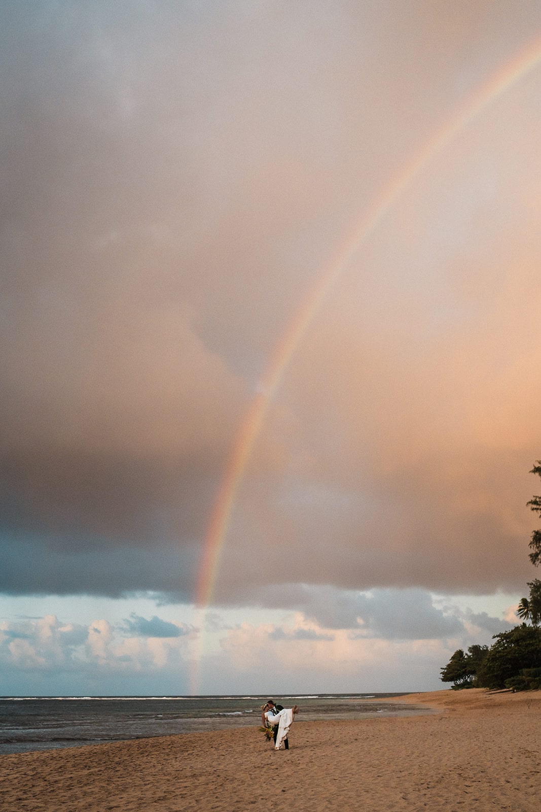 Rainbow fills the sky while groom dips bride for a kiss on the beach during their Kauai micro wedding weekend.