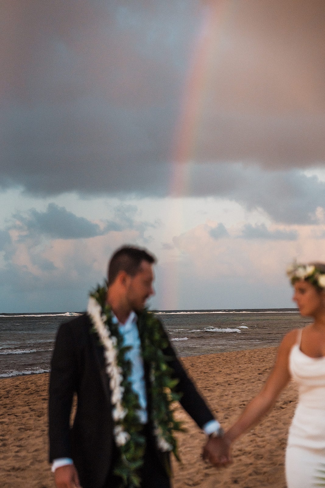 Bride and groom hold hands on the beach while a rainbow fills the sky behind them at their Kauai wedding.
