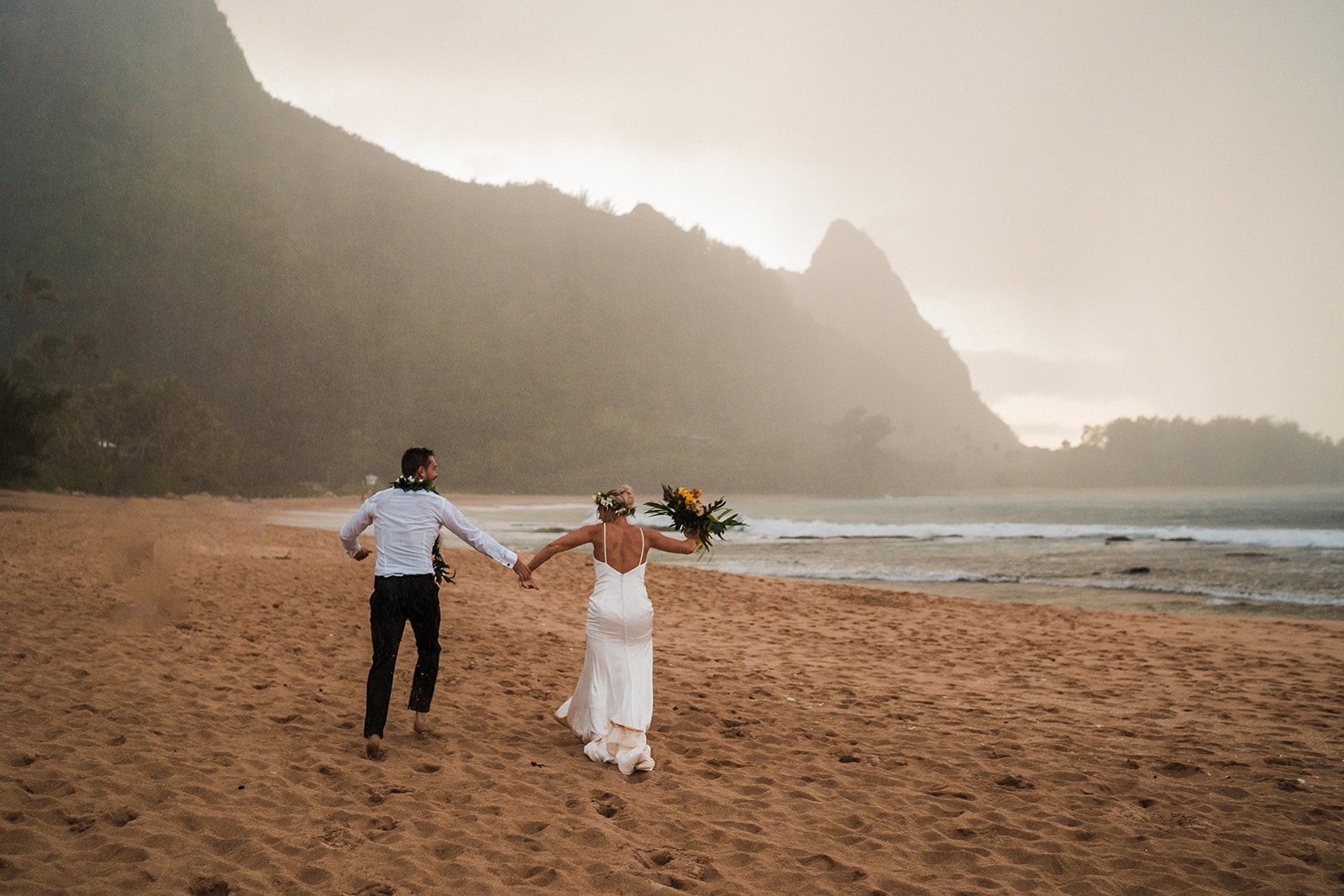 Bride and groom hold hands while running across a Kauai beach in the rain.