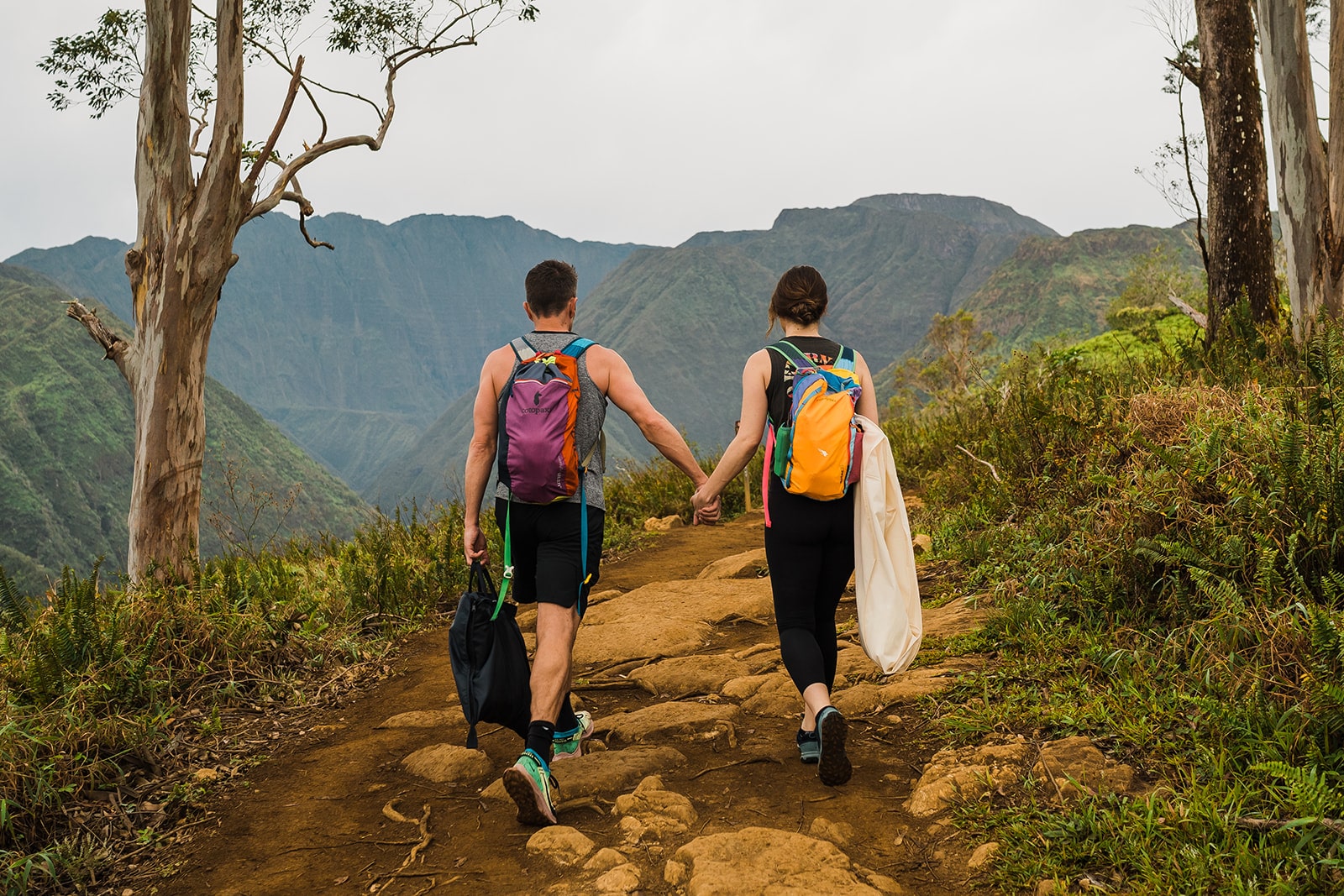 Bride and groom hold hands while walking down a red rock trail and carrying their wedding attire to their ceremony location for their elopement in Kauai.