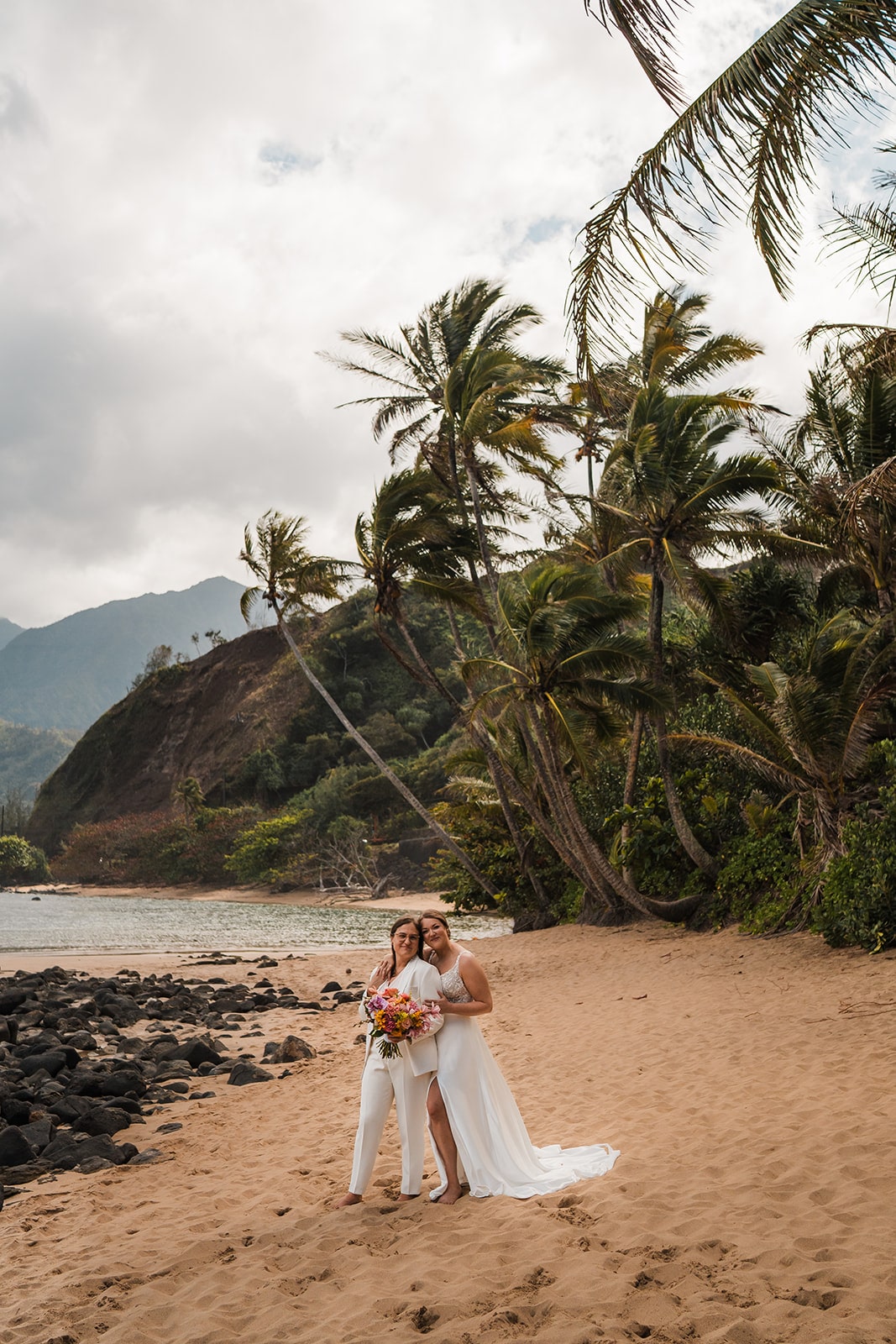 Two brides stand side by side at one of the best Kauai elopement locations on a beach.