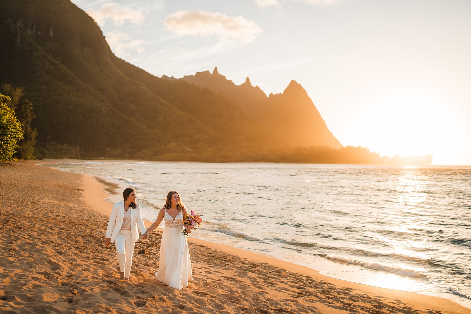 Two brides hold hands while walking across the beach at sunset during their elopement in Kauai.