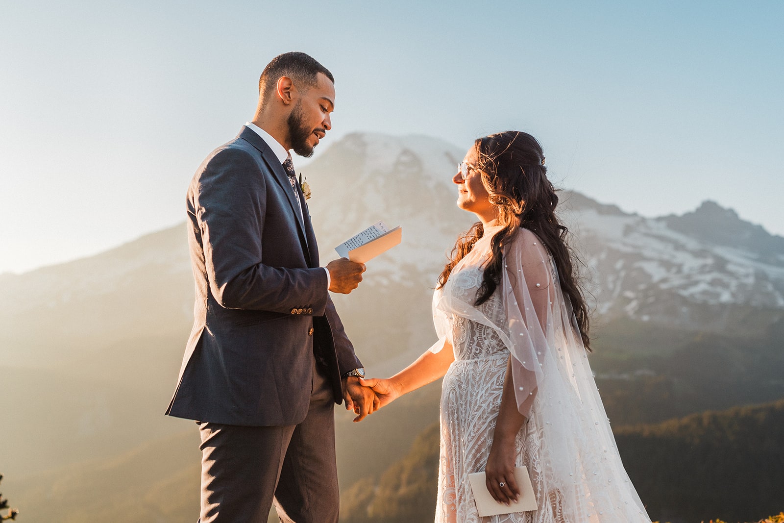 Groom reads vows while holding bride's hand on a mountain trail during their Mount Rainier National Park elopement.