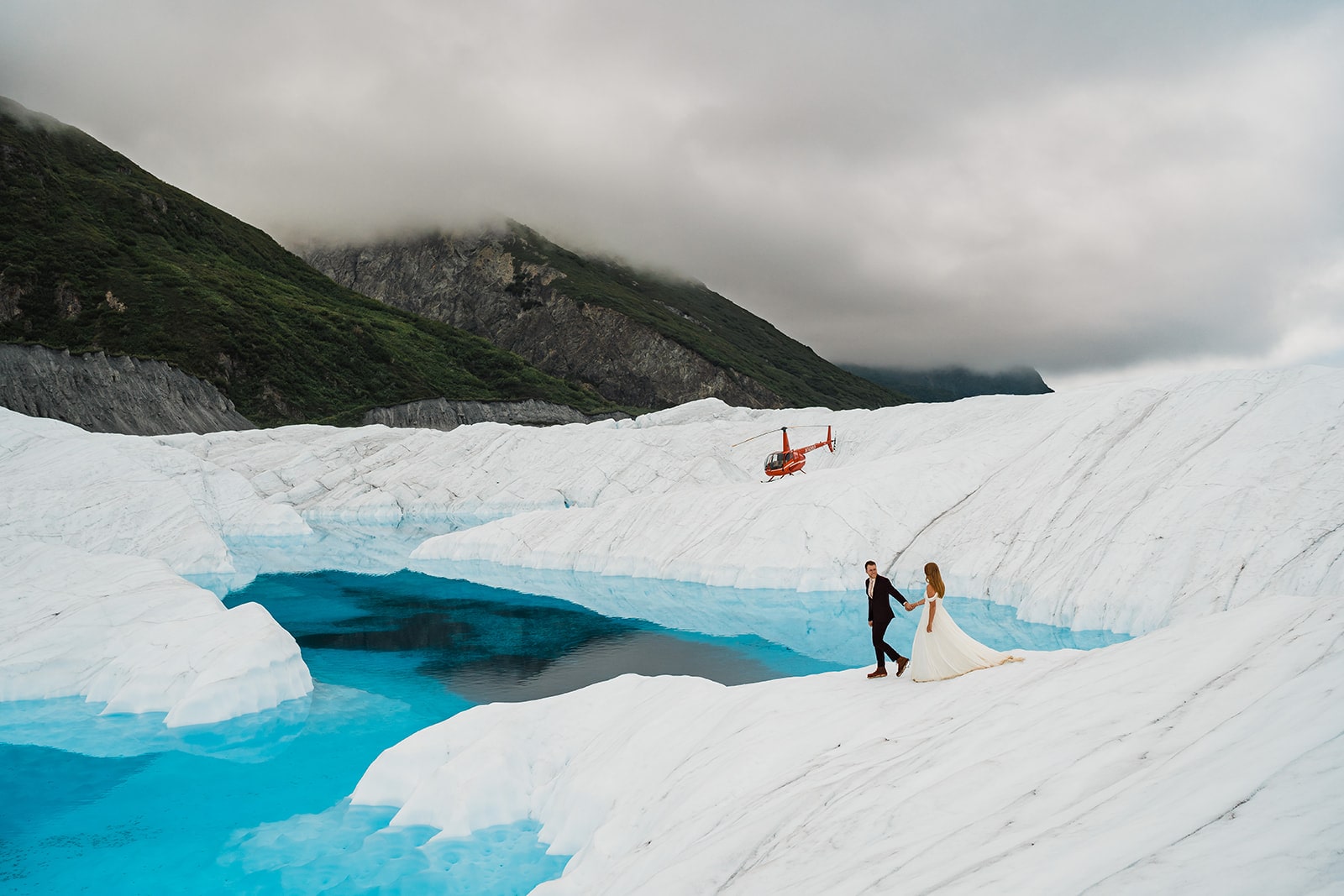 Bride and groom hold hands as they walk across an icy glacier in Alaska during their helicopter elopement.
