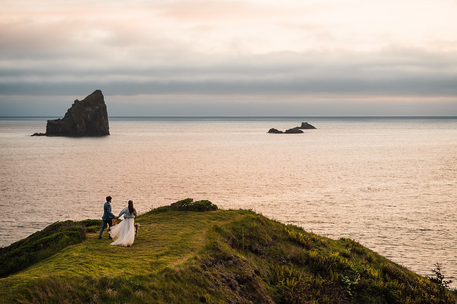 Bride and groom hold hands while walking out to the edge of the cliffs on the Oregon Coast during their elopement at sunset.