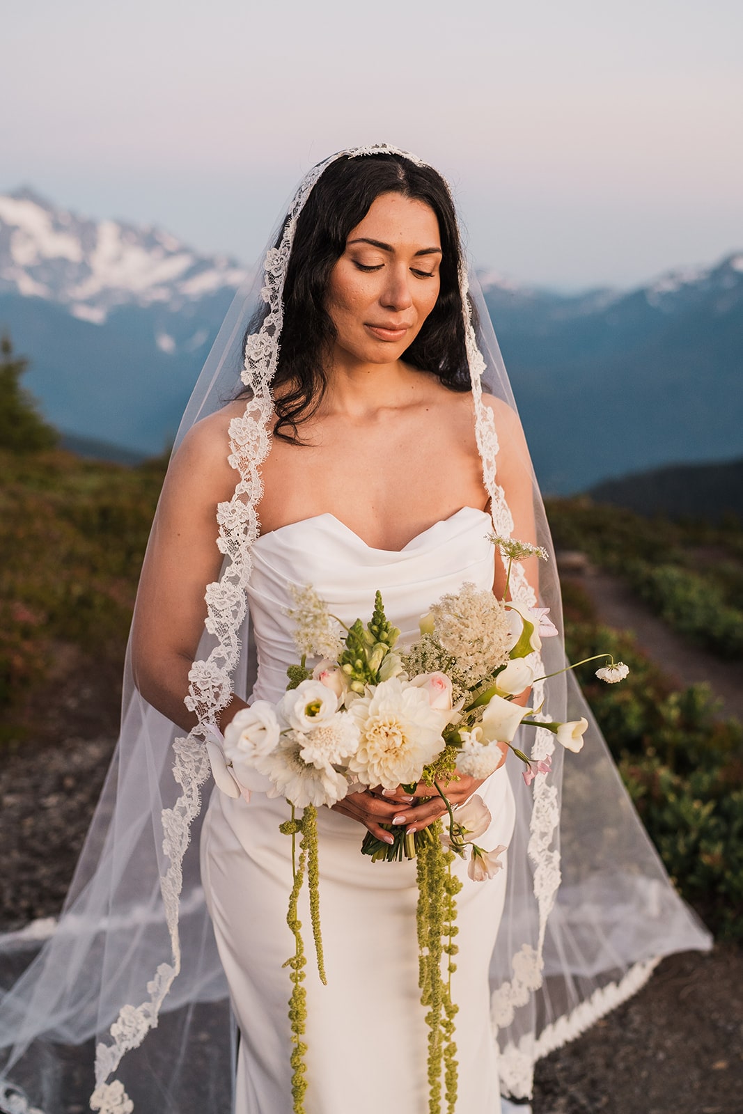 Bride wearing white wedding dress with lace trimmed veil holds a trailing flower bouquet in the North Cascade mountains.